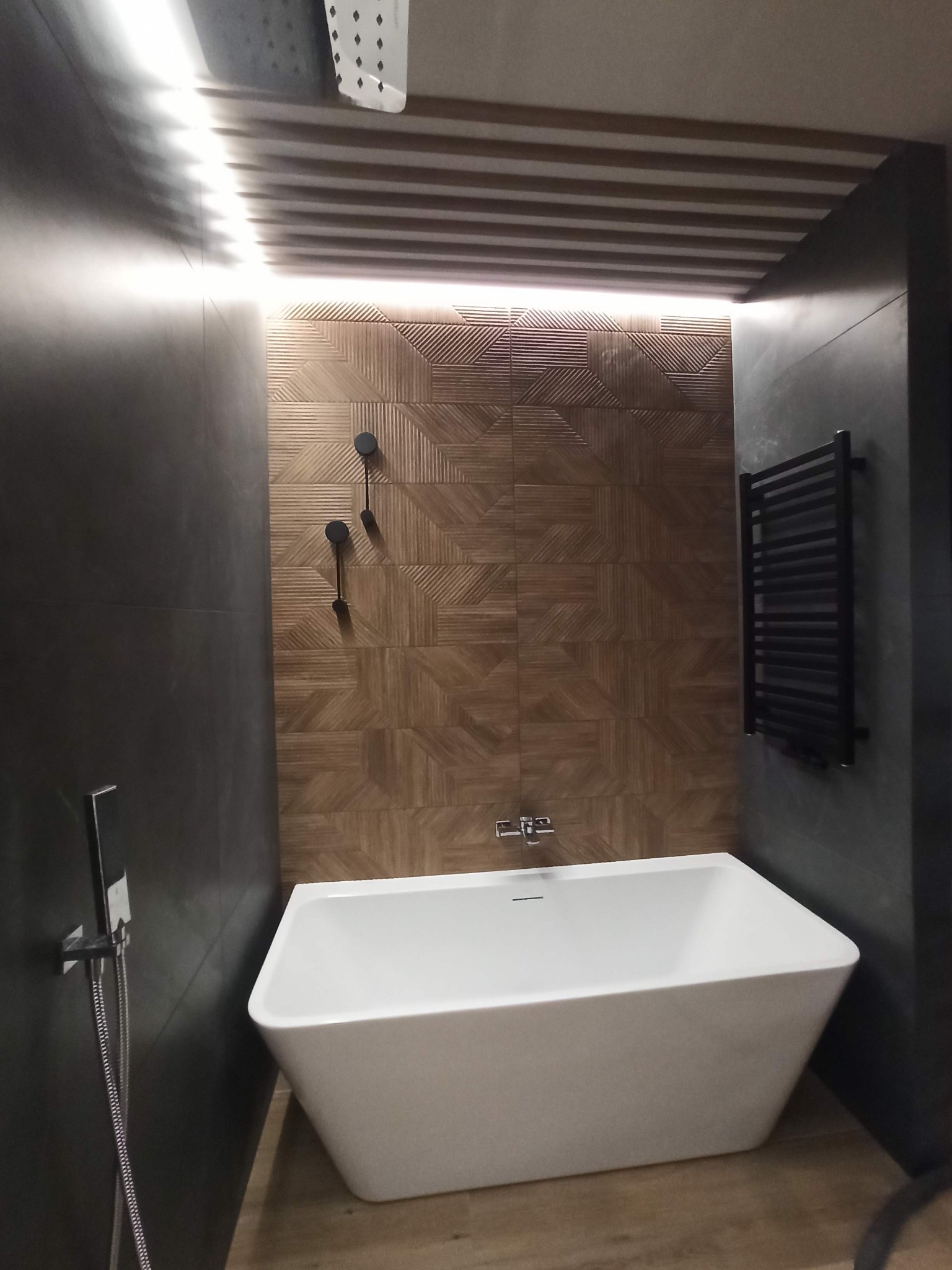 Nowoczesna łazienka z wolnostojącą białą wanną, czarnymi płytkami na ścianach, drewnopodobną płytką dekoracyjną na ścianie i suficie z podświetleniem LED.