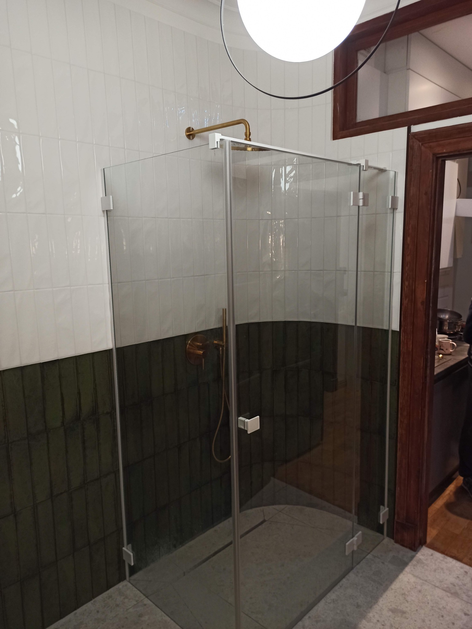 Szklana kabina prysznicowa z złotą armaturą na tle dwukolorowych płytek w łazience z drewnianą ramą drzwiową