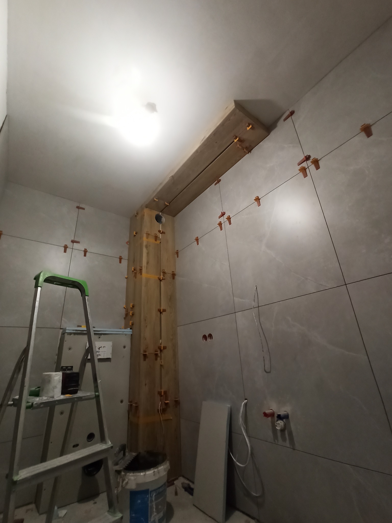 Montaż szarych płytek w łazience, widoczne krzyżyki dystansowe, drewniana konstrukcja pod sufitem, drabina z narzędziami i resztkami kleju, przygotowane rury do podłączenia.