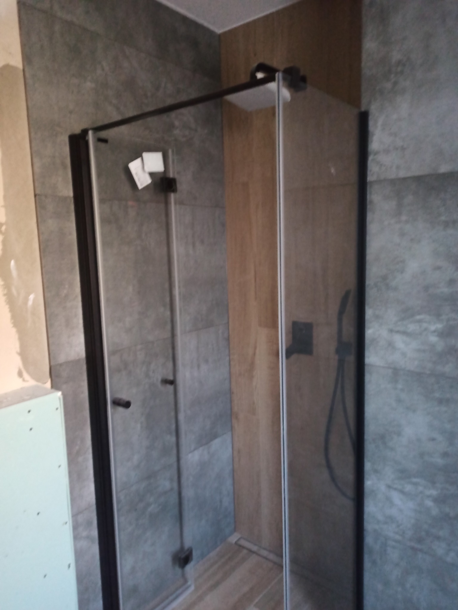 Nowoczesna kabina prysznicowa z czarnymi profilami, zamontowana w łazience wykończonej płytkami w odcieniach szarości i drewna, widoczne elementy instalacji prysznicowej.