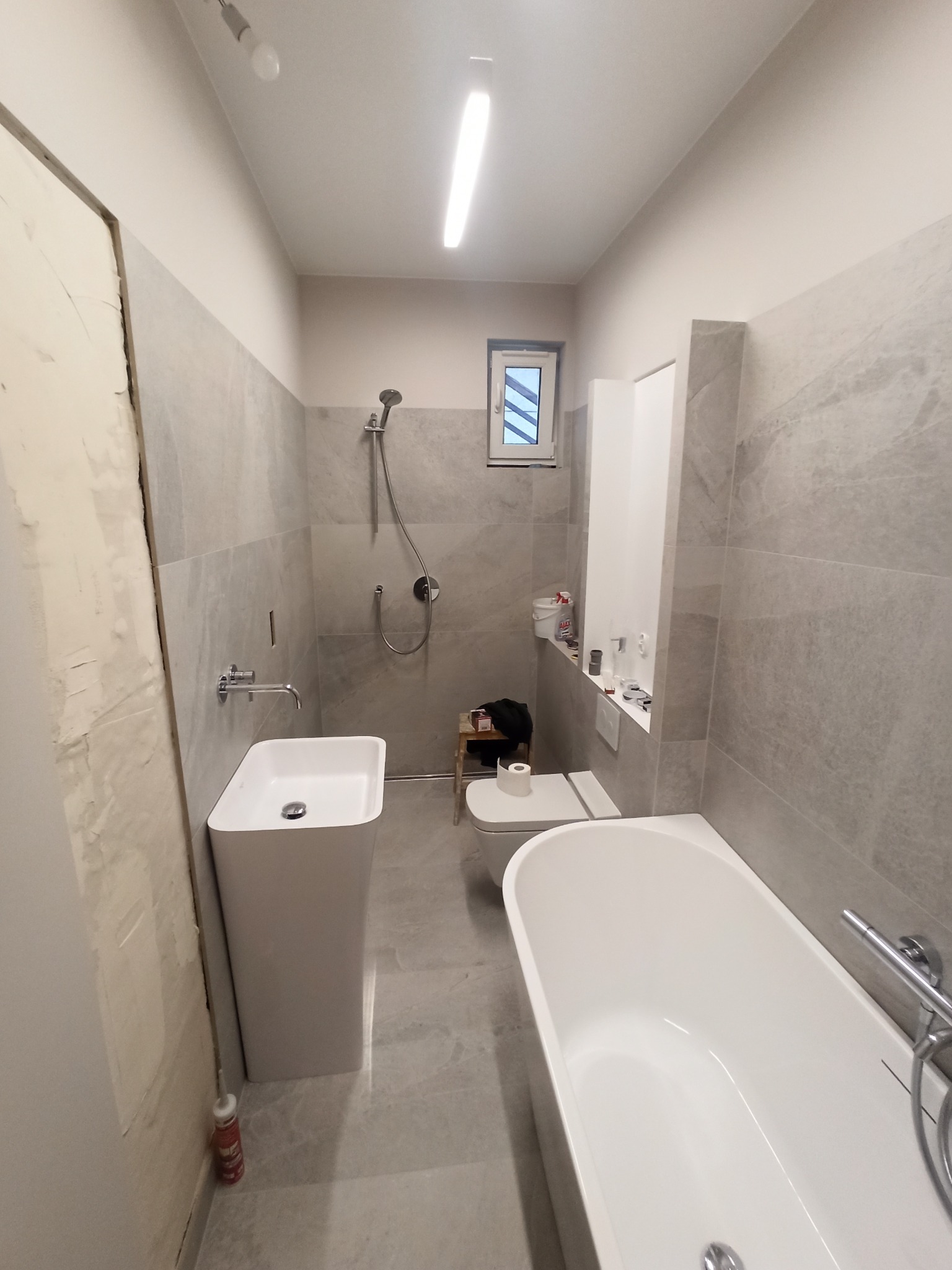 Łazienka w trakcie remontu z wanną, umywalką na postumencie, toaletą i szarymi płytkami imitującymi marmur; widoczne niedokończone wykończenia przy drzwiach.