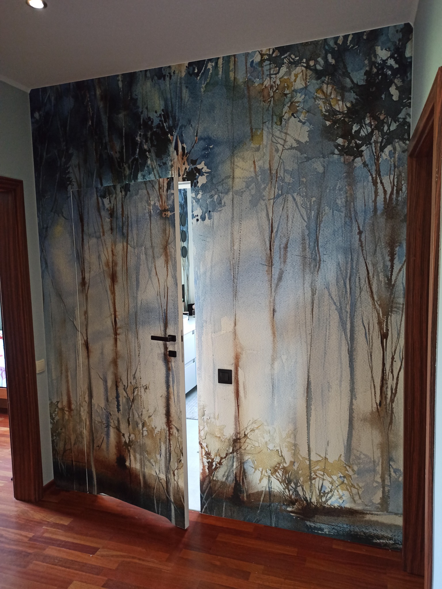 Drzwi przesuwne z fototapetą imitującą akwarelowy las, zamontowane w pokoju z parkietem w odcieniach czerwieni.