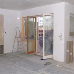 Wnętrze w trakcie remontu: surowe ściany, częściowo położona gładź, otwarte białe drzwi balkonowe z drewnianą ramą, drabina, rolki papieru zabezpieczającego podłogę.