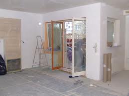 Wnętrze w trakcie remontu: surowe ściany, częściowo położona gładź, otwarte białe drzwi balkonowe z drewnianą ramą, drabina, rolki papieru zabezpieczającego podłogę.