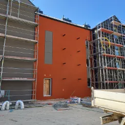 Budynek wielorodzinny w trakcie prac elewacyjnych: ocieplanie i tynkowanie, widoczne rusztowania, materiały budowlane i fragment skrzyni ładunkowej samochodu.