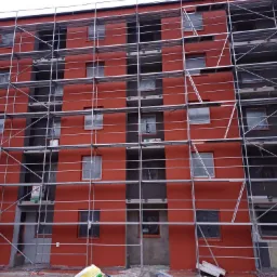 Czerwona fasada budynku wielopiętrowego w trakcie prac wykończeniowych, z widocznymi rusztowaniami i oknami zabezpieczonymi folią.