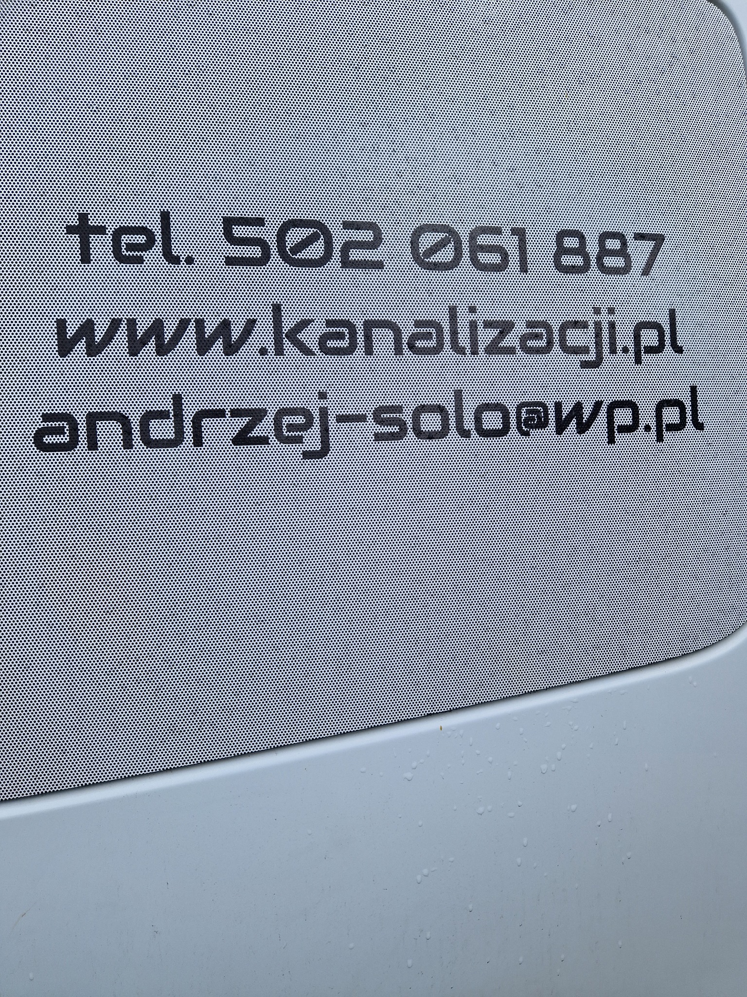 Reklama firmy kanalizacyjnej z numerem telefonu, adresem strony internetowej i adresem e-mail umieszczona na perforowanej folii okiennej na samochodzie.