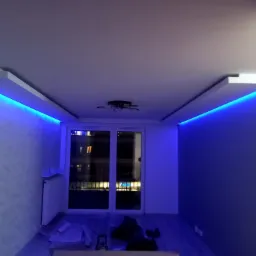 Pokój z oświetleniem LED w listwach sufitowych, świecących na niebiesko, widoczne okno balkonowe z widokiem na nocne miasto, na podłodze folie malarskie.