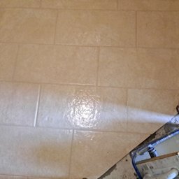 Mateusz Węgielnik - Układanie beżowych, prostokątnych płytek na podłodze, widoczny fragment narzędzia do rozprowadzania fugi i świeżo nałożona fuga na płytkach.