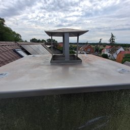 Mateusz Węgielnik - Metalowy komin z daszkiem na tle dachu z panelami słonecznymi i zabudowy wiejskiej pod pochmurnym niebem.