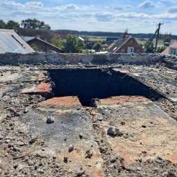 Mateusz Węgielnik - Otwór w zniszczonym, ceglanym kominie na dachu budynku z widokiem na okoliczne domy i pola w słoneczny dzień.