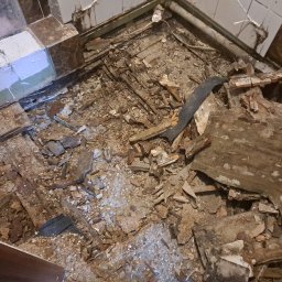 Mateusz Węgielnik - Zdemontowana podłoga w łazience, widoczne fragmenty desek, gruz i stare płytki na ścianach.