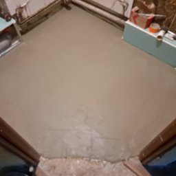 Mateusz Węgielnik - Wylany, świeży podkład podłogowy w łazience, widoczne rury, kartony i narzędzia na ścianie, surowy stan wykończenia.