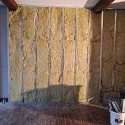 Ściana wewnętrzna w trakcie izolacji wełną mineralną, widoczne metalowe profile, drewniane belki stropowe i fragment kominka z boku