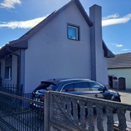 Mateusz Węgielnik - Otynkowany dom jednorodzinny z szarą elewacją i ciemnym dachem, widoczny zza metalowego ogrodzenia, z zaparkowanym czarnym samochodem osobowym i fragmentem białego samochodu dostawczego po prawej...