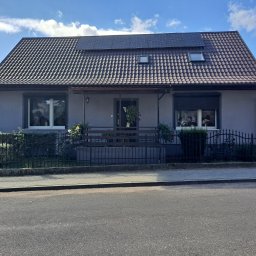 Mateusz Węgielnik - Dom jednorodzinny w Gorzowie Wielkopolskim z szarą elewacją, brązowym dachem pokrytym panelami słonecznymi, oraz niewielkim zadaszonym tarasem z czarną balustradą, otoczony zielenią.