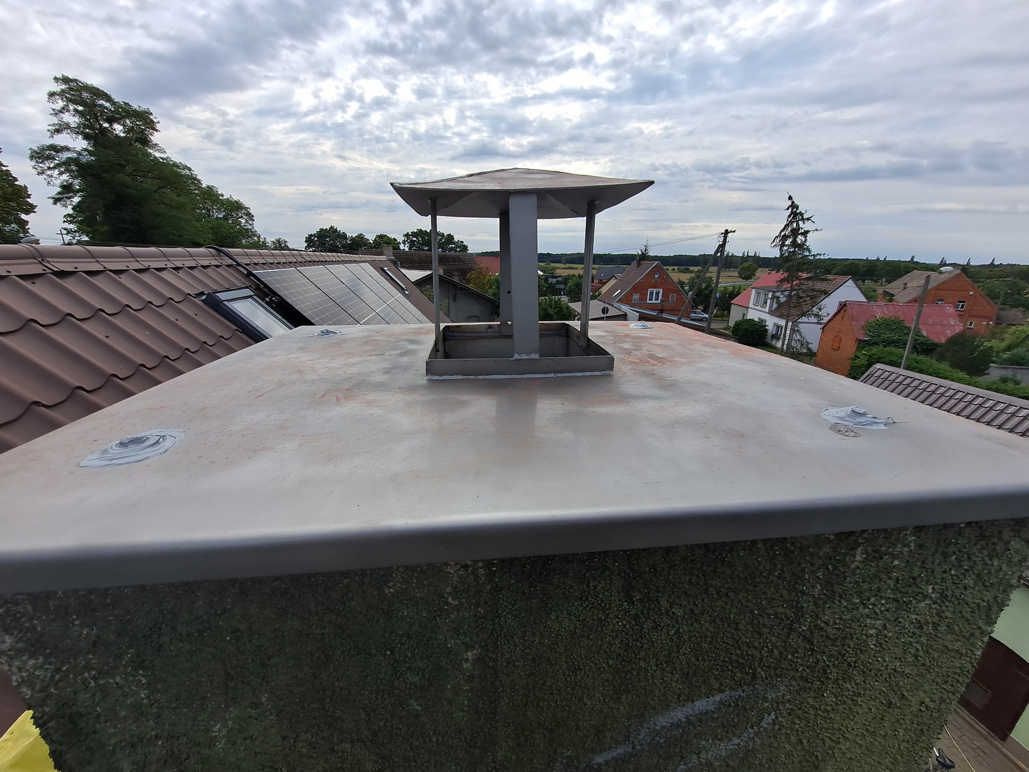 Metalowy komin z daszkiem na tle dachu z panelami słonecznymi i zabudowy wiejskiej pod pochmurnym niebem.