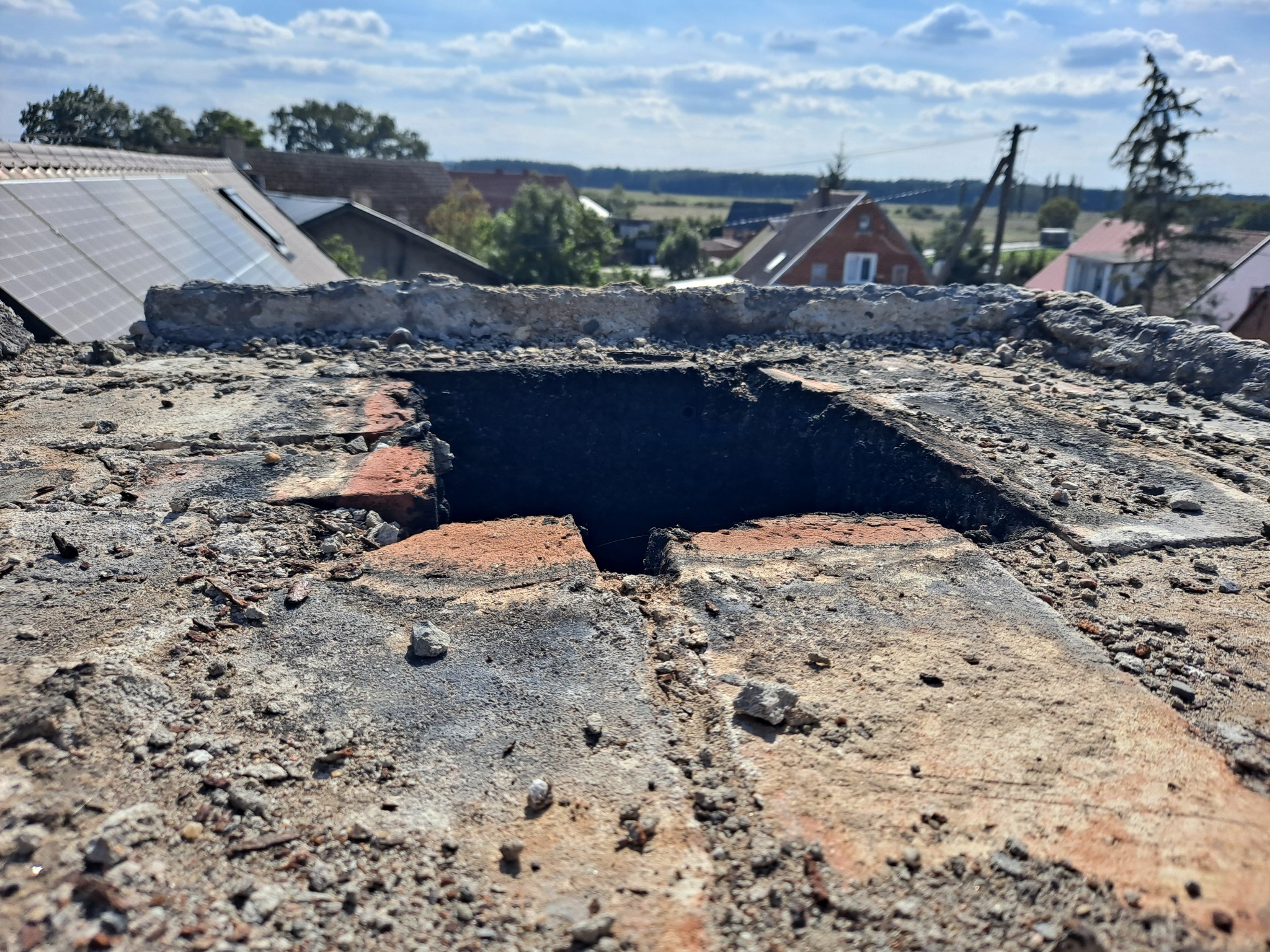 Otwór w zniszczonym, ceglanym kominie na dachu budynku z widokiem na okoliczne domy i pola w słoneczny dzień.