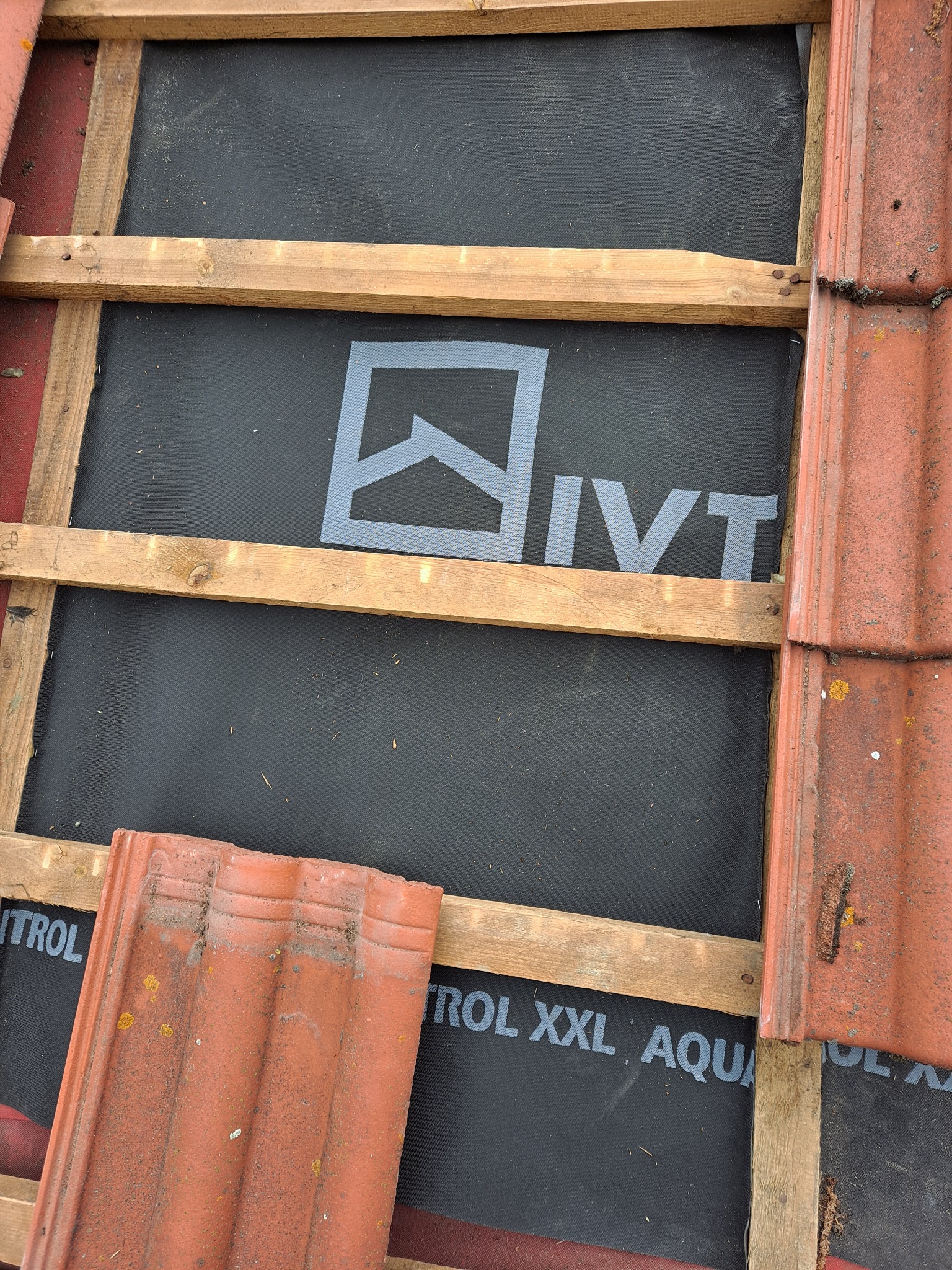 Fragment dachu z widoczną membraną dachową z nadrukiem, drewnianą konstrukcją nośną i ułożonymi dachówkami ceramicznymi w odcieniach czerwieni, częściowo zabrudzonymi mchem.