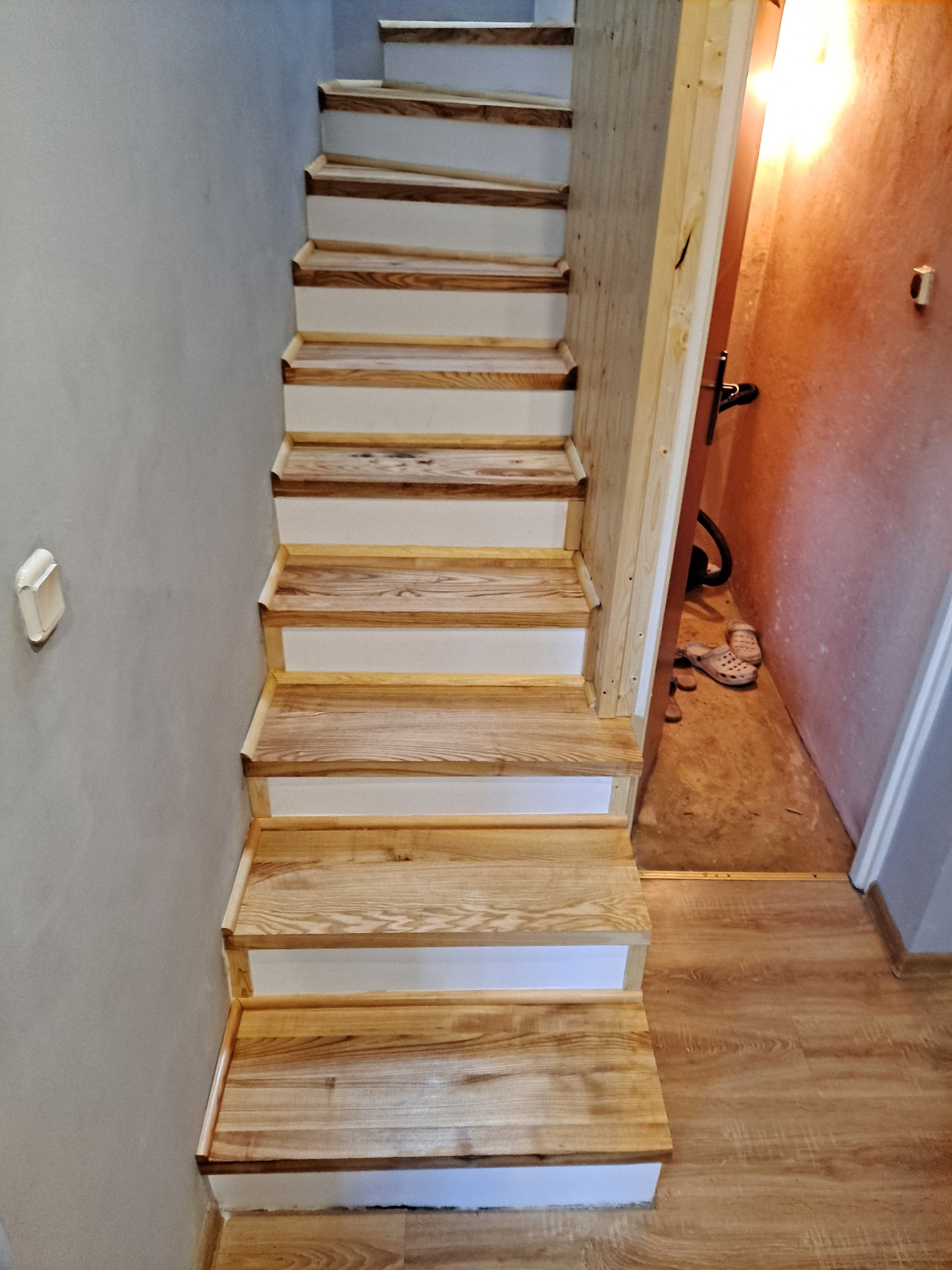Drewniane schody z białymi podstopnicami w trakcie montażu, widoczne niedoskonałości wykończenia i robocze obuwie w przejściu.