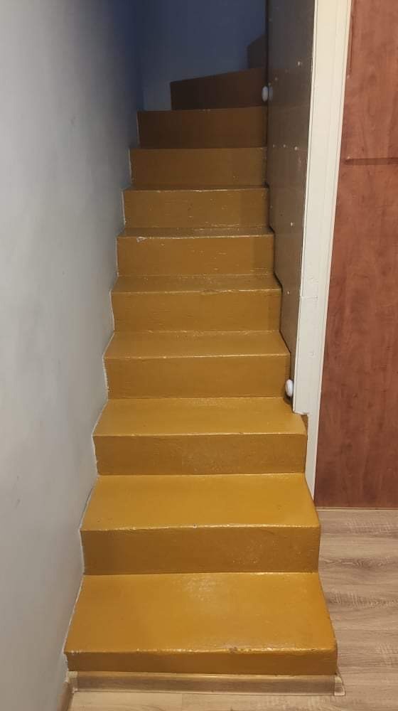Schody wewnętrzne pomalowane na kolor musztardowy, widoczne ślady niedokładnego malowania przy ścianie i drzwiach.