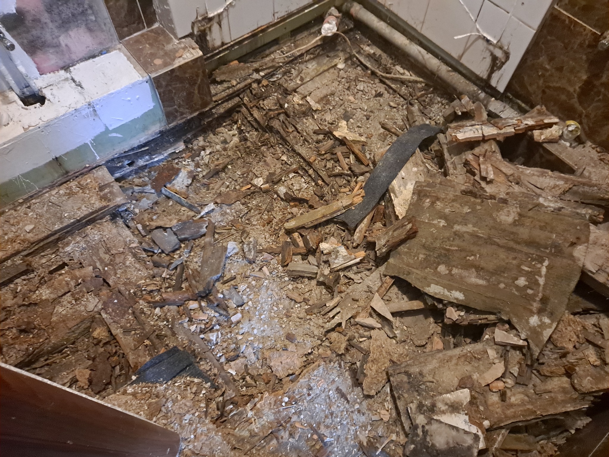 Zdemontowana podłoga w łazience, widoczne fragmenty desek, gruz i stare płytki na ścianach.