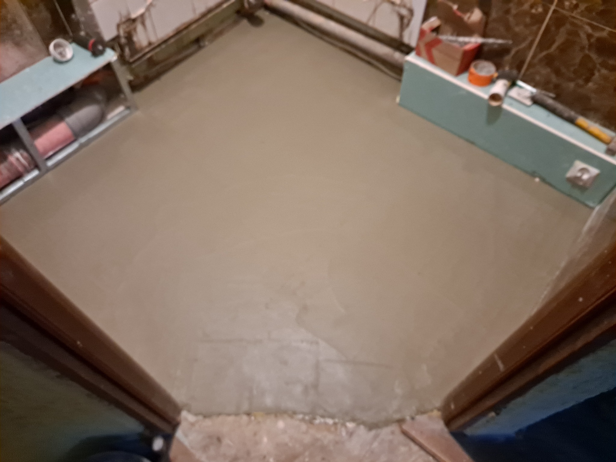 Wylany, świeży podkład podłogowy w łazience, widoczne rury, kartony i narzędzia na ścianie, surowy stan wykończenia.