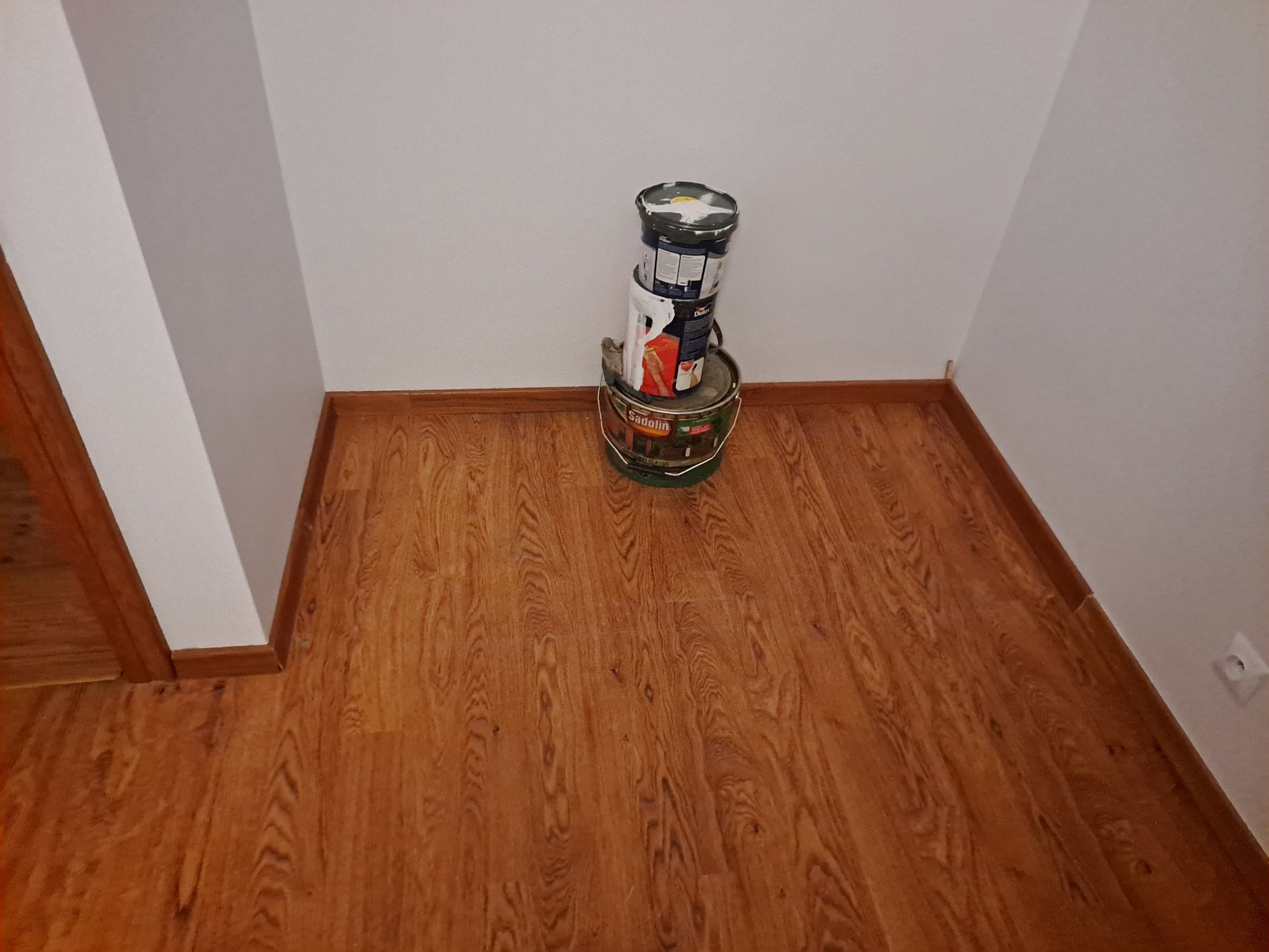 Stojące jedna na drugiej puszki z farbą różnych marek, ustawione w rogu pokoju na drewnianej podłodze przy białej ścianie z listwą przypodłogową.