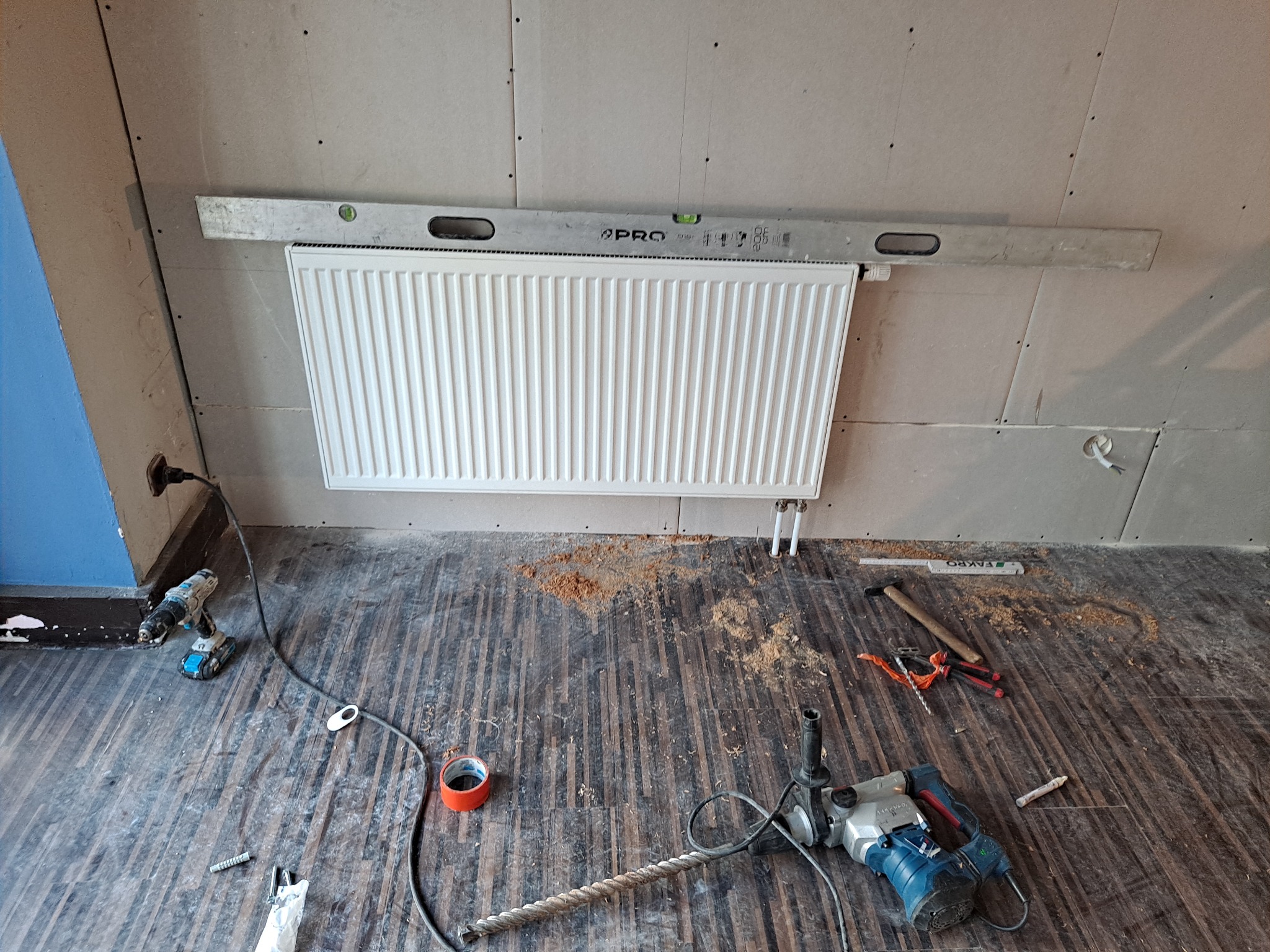 Montaż grzejnika na ścianie z płyt kartonowo-gipsowych, poziomica przymocowana nad grzejnikiem, wiertarka udarowa i wkrętarka na podłodze z paneli, widoczne ślady po wierceniu.