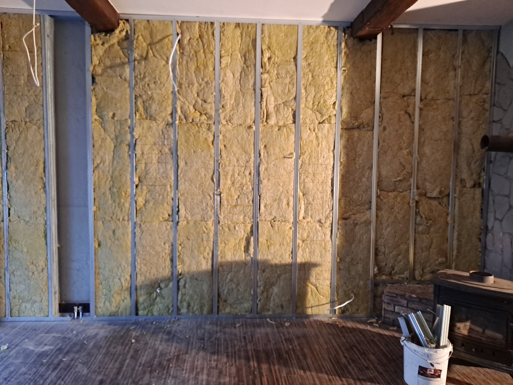 Ściana wewnętrzna w trakcie izolacji wełną mineralną, widoczne metalowe profile, drewniane belki stropowe i fragment kominka z boku