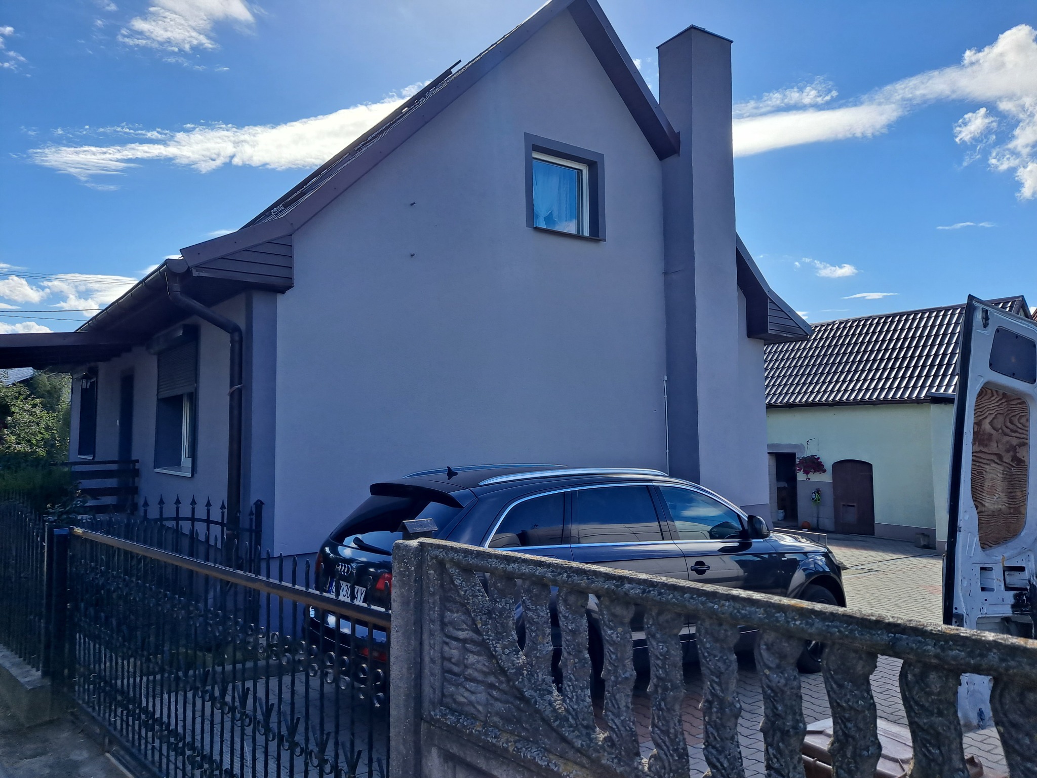 Otynkowany dom jednorodzinny z szarą elewacją i ciemnym dachem, widoczny zza metalowego ogrodzenia, z zaparkowanym czarnym samochodem osobowym i fragmentem białego samochodu dostawczego po prawej...