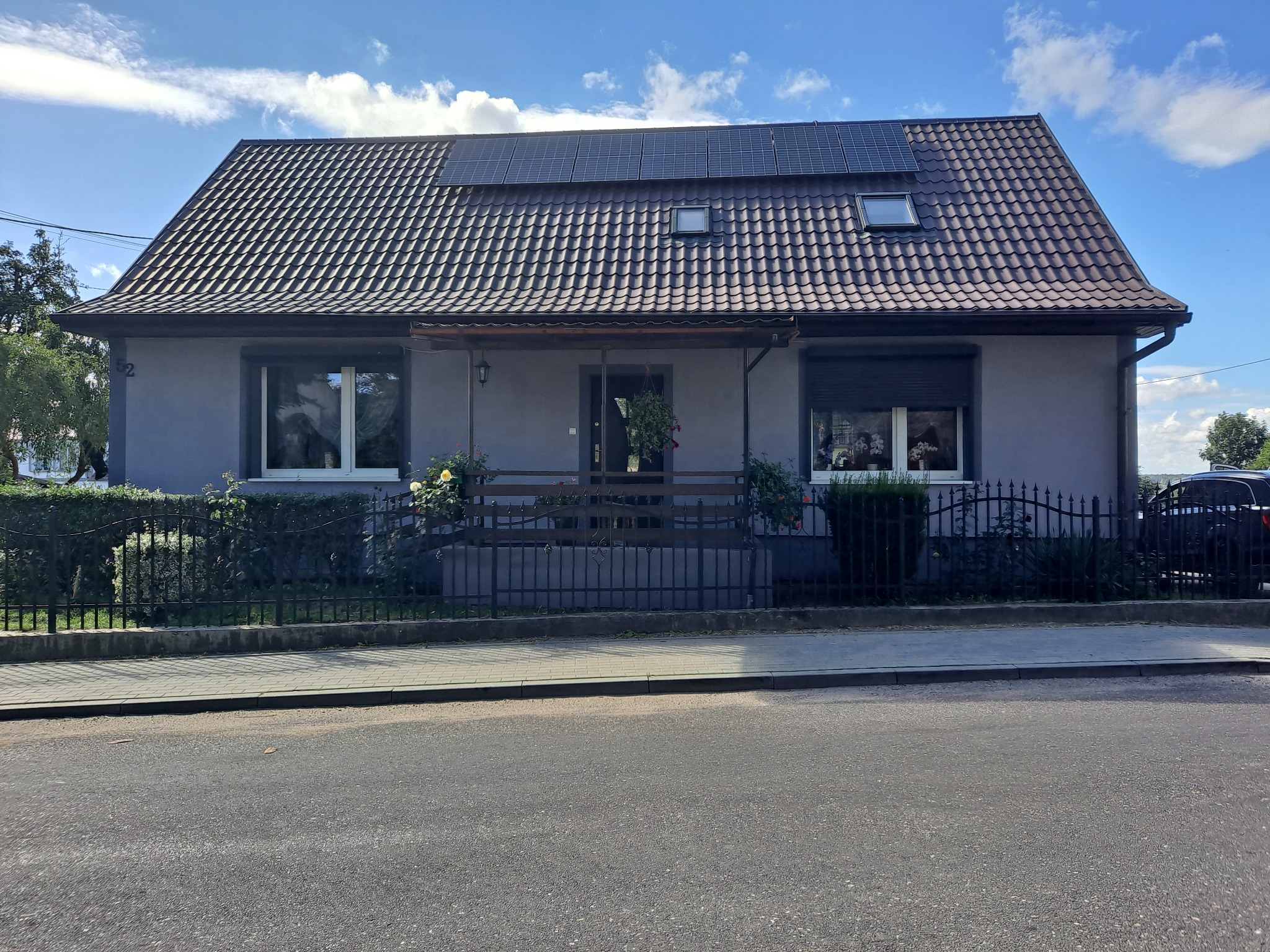 Dom jednorodzinny w Gorzowie Wielkopolskim z szarą elewacją, brązowym dachem pokrytym panelami słonecznymi, oraz niewielkim zadaszonym tarasem z czarną balustradą, otoczony zielenią.