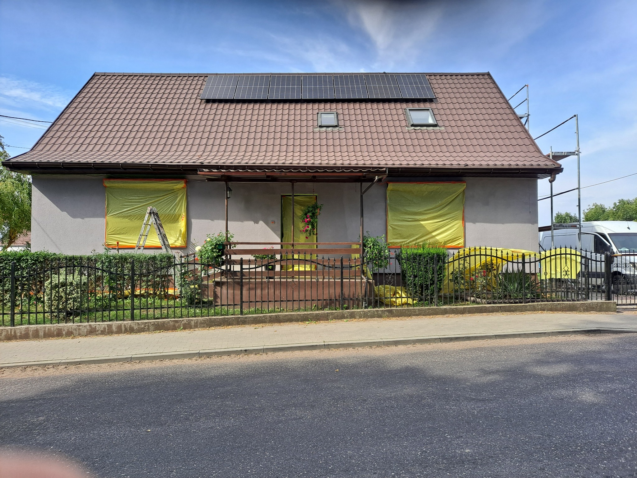 Dom jednorodzinny z panelami słonecznymi na dachu, okna i drzwi zabezpieczone żółtą folią ochronną, rusztowanie przy ścianie bocznej, widok z ulicy.