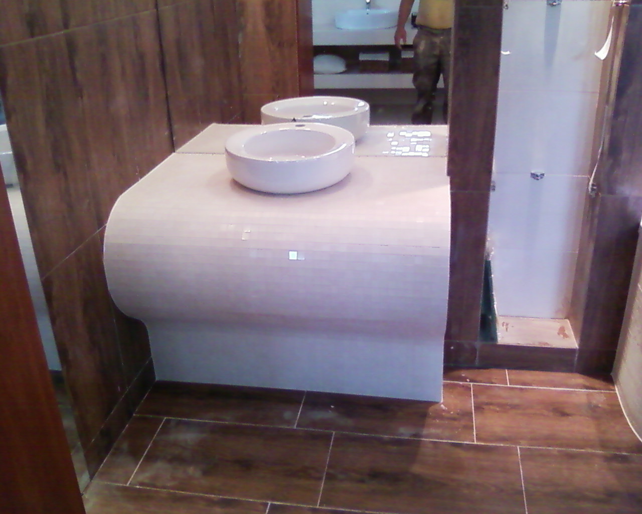 Umywalka nablatowa w kształcie misy na podmurowanym blacie wykończonym mozaiką, drewnopodobna glazura na ścianach i podłodze, widoczne odbicie osoby w lustrze.