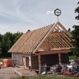 Budowa domu jednorodzinnego w trakcie realizacji, widoczna konstrukcja dachu z wieńcem, częściowo otynkowane ściany, materiały budowlane na placu budowy.