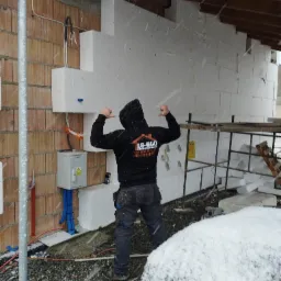 Pracownik w czarnej bluzie z logo firmy ES-BUD, z podniesionymi rękami, prezentuje fragment ocieplonej ściany budynku w trakcie śnieżycy. Widoczna ceglana ściana, białe płyty izolacyjne, skrzynka...