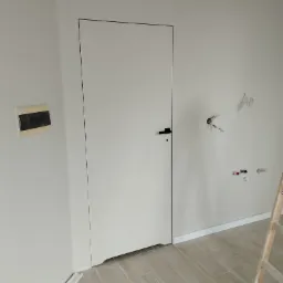 Świeżo pomalowane, jasne wnętrze mieszkania z ukrytymi drzwiami z czarną klamką, widoczne niedokończone instalacje elektryczne i hydrauliczne, oraz fragment drabiny malarskiej.