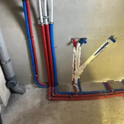 Surowe wnętrze w trakcie prac hydraulicznych: widoczne rury doprowadzające wodę w kolorach czerwonym i niebieskim, częściowo zamocowane do ściany, z widocznymi otworami w ścianie i pianką montażową...