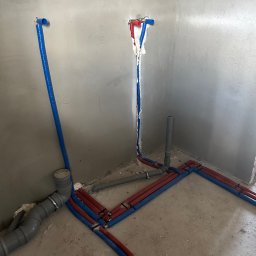 Marek Thrun - Surowe wnętrze w trakcie prac hydraulicznych: rury wodne (czerwone i niebieskie) prowadzone wzdłuż ścian i podłogi, pionowe wyprowadzenia ze ściany zabezpieczone pianką montażową, widoczne elementy...