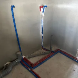 Surowe wnętrze w trakcie prac hydraulicznych: rury wodne (czerwone i niebieskie) prowadzone wzdłuż ścian i podłogi, pionowe wyprowadzenia ze ściany zabezpieczone pianką montażową, widoczne elementy...