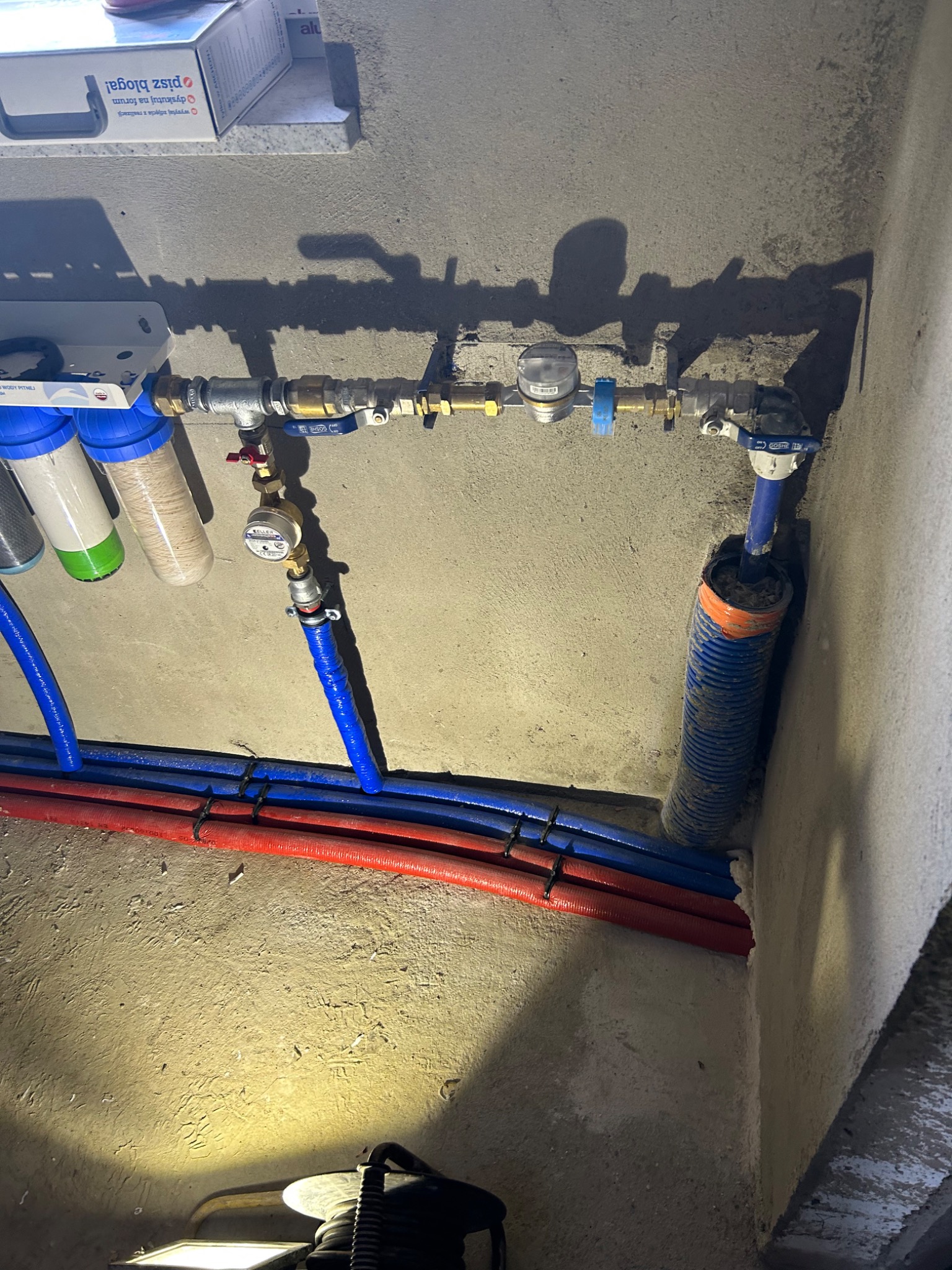 Instalacja wodna z filtrami, licznikiem i rurami w kolorach niebieskim i czerwonym, zamocowana na szarej ścianie. Rury zabezpieczone czarnymi opaskami zaciskowymi. Część rury w niebieskiej...