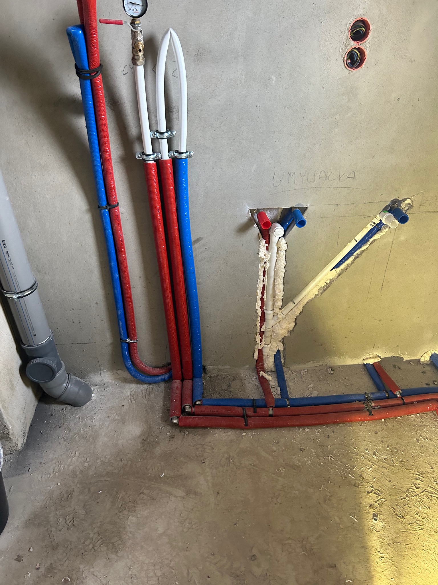 Surowe wnętrze w trakcie prac hydraulicznych: widoczne rury doprowadzające wodę w kolorach czerwonym i niebieskim, częściowo zamocowane do ściany, z widocznymi otworami w ścianie i pianką montażową...