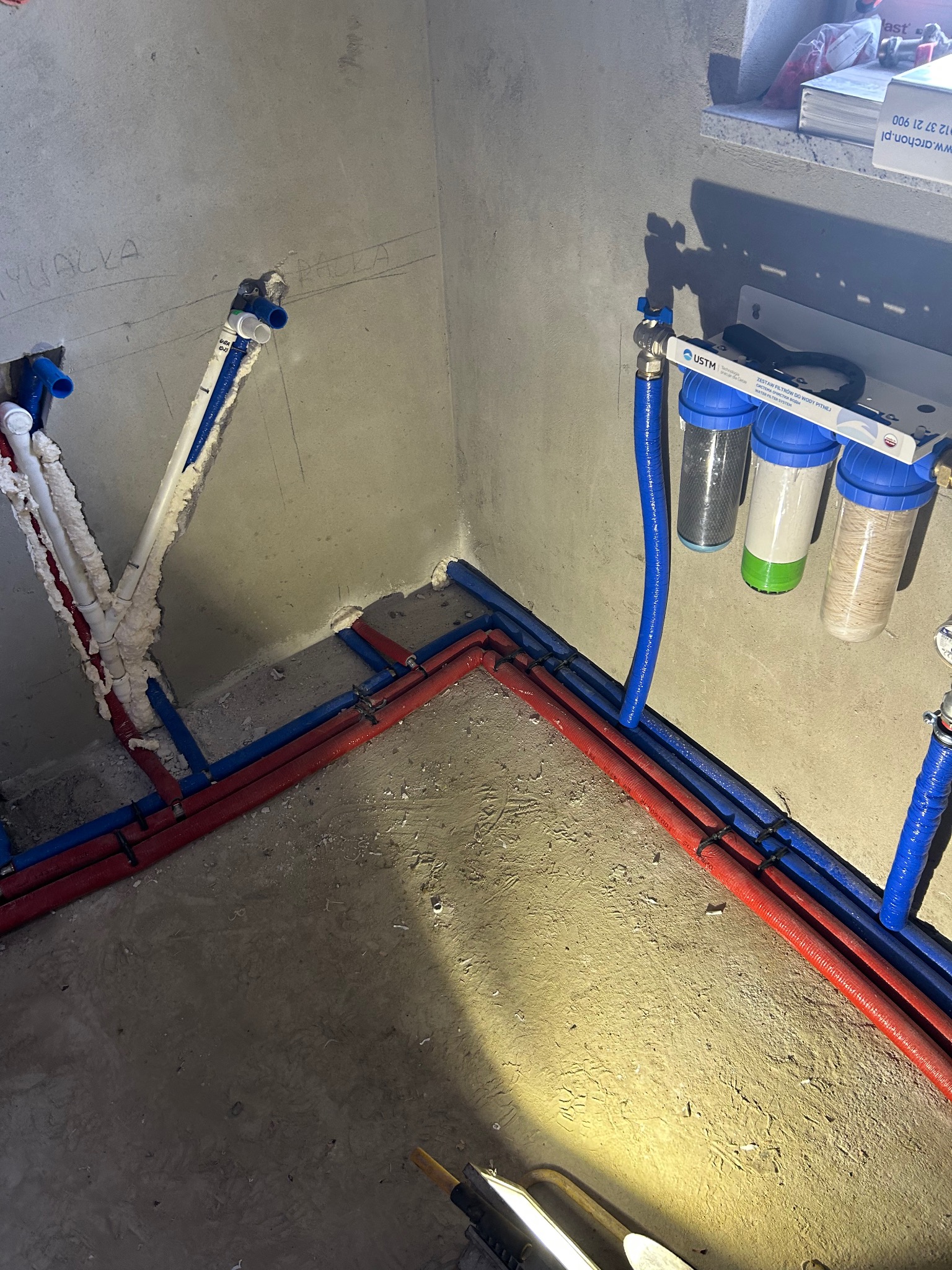 Montaż instalacji wodnej w surowym pomieszczeniu: widoczne czerwone i niebieskie rury prowadzone po podłodze i ścianach, zabezpieczone pianką montażową, oraz zestaw filtrów do wody pitnej USTM...