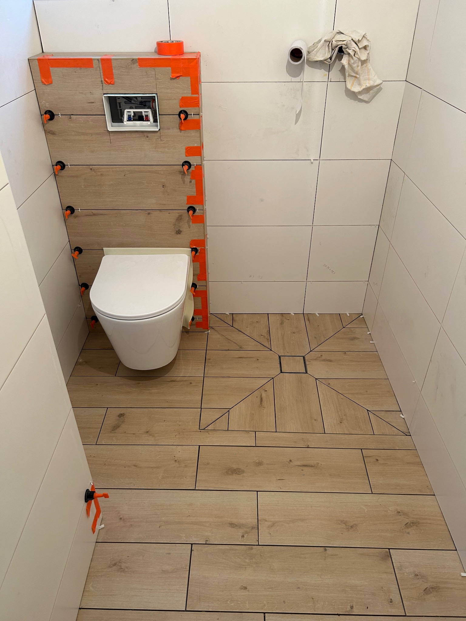 Montaż podwieszanej toalety z deską sedesową w łazience z drewnopodobnymi płytkami, widoczne pomarańczowe krzyżyki dystansowe i taśma maskująca na obudowie stelaża podtynkowego, częściowo ułożona...