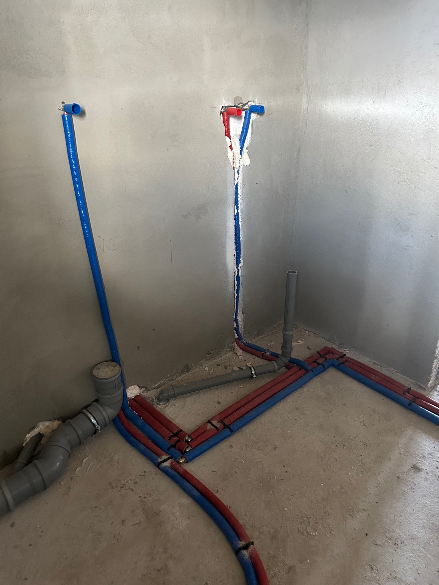 Surowe wnętrze w trakcie prac hydraulicznych: rury wodne (czerwone i niebieskie) prowadzone wzdłuż ścian i podłogi, pionowe wyprowadzenia ze ściany zabezpieczone pianką montażową, widoczne elementy...