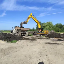 Żółta koparka JCB JS200 nabiera ziemię i wysypuje ją do wywrotki na tle błękitnego nieba z delikatnymi chmurami.