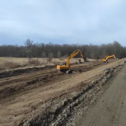 Żółte koparki pracują na placu budowy, przygotowując teren pod drogę; w tle widoczny las bez liści.