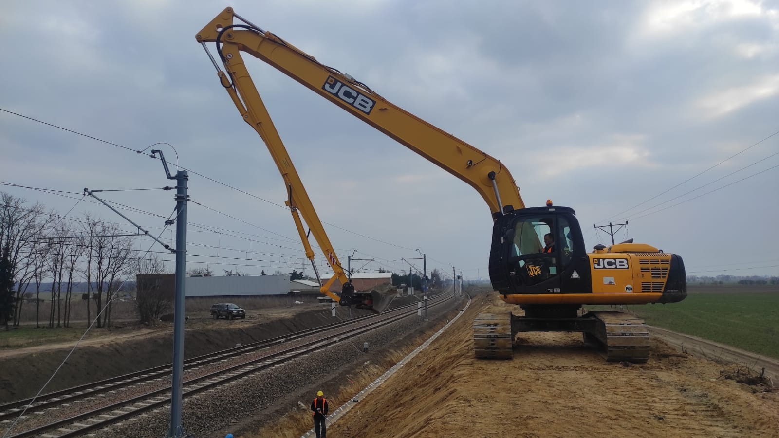 Żółta koparka JCB przy torach kolejowych, wyrównująca teren w pobliżu słupa trakcyjnego, pracownik w kasku na dole kadru, pochmurne niebo.