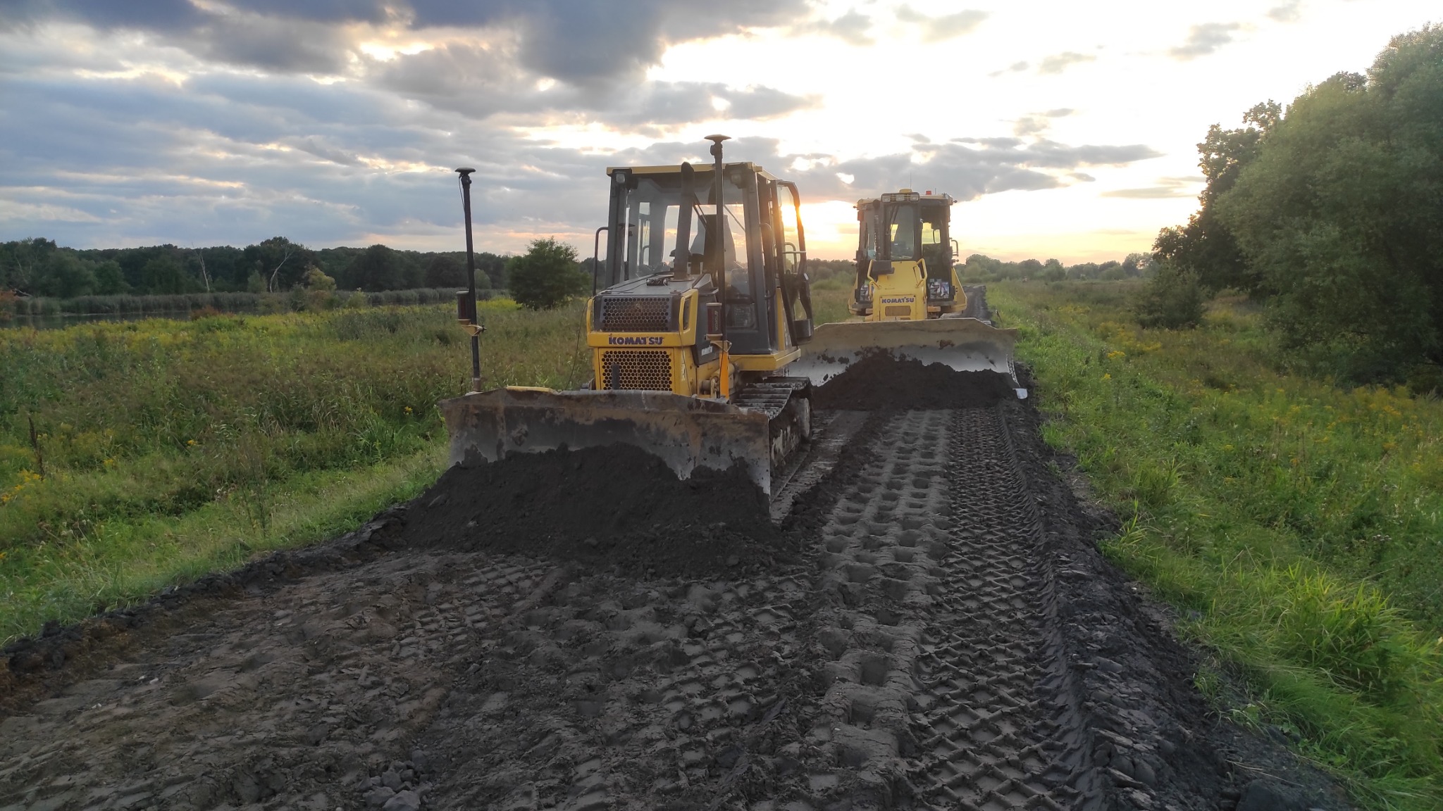 Dwa spychacze Komatsu pracują na polu, wyrównując ziemię pod budowę drogi, widoczne ślady gąsienic na ciemnej glebie, w tle zachodzące słońce i zielona roślinność.