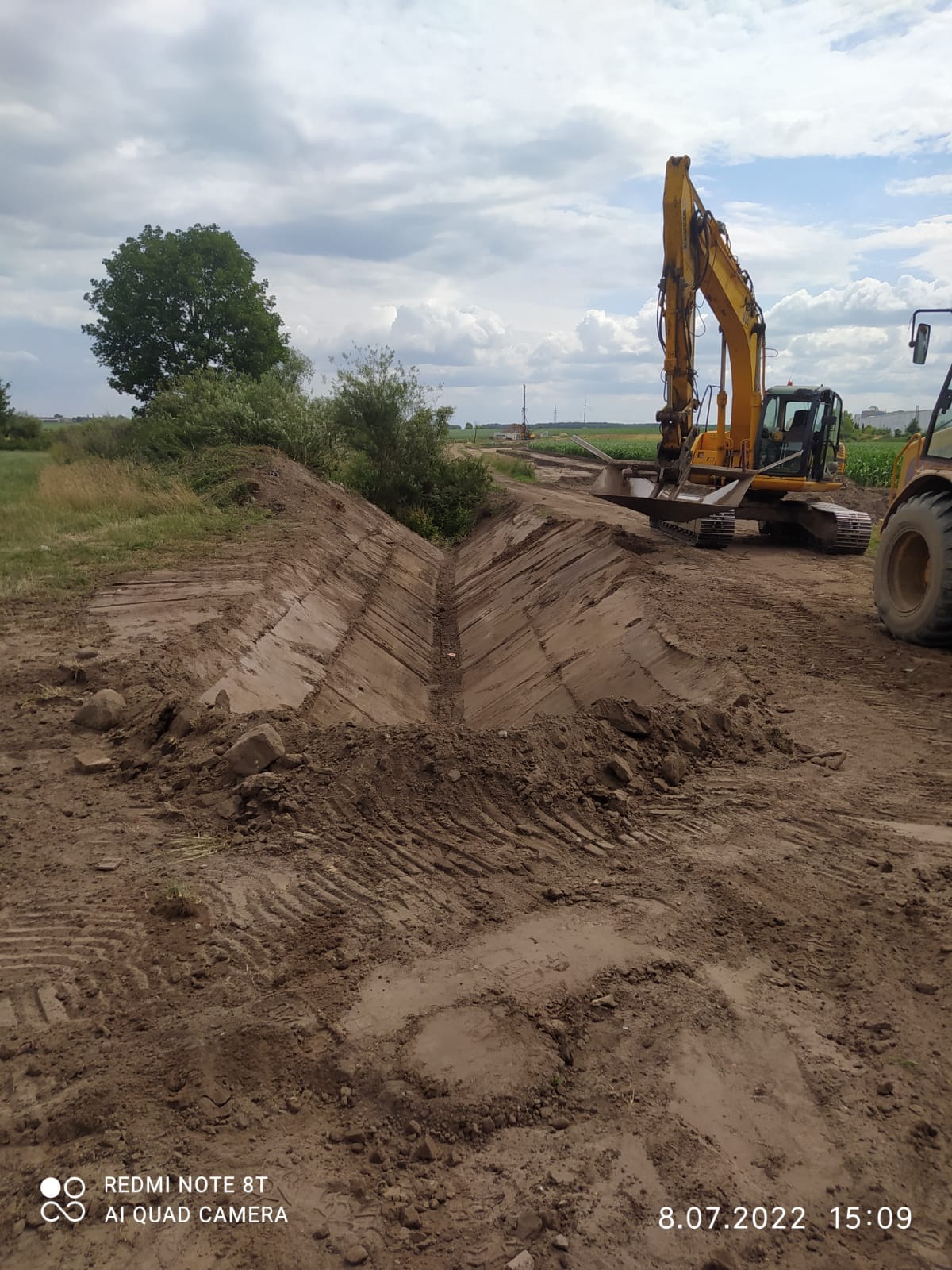 Żółta koparka gąsienicowa obok wykopanego rowu melioracyjnego z widocznymi śladami gąsienic i opon, w tle drzewa i pochmurne niebo.